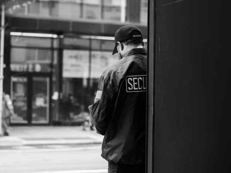 A security guard in a dark jacket stands in a doorway, seen from behind.